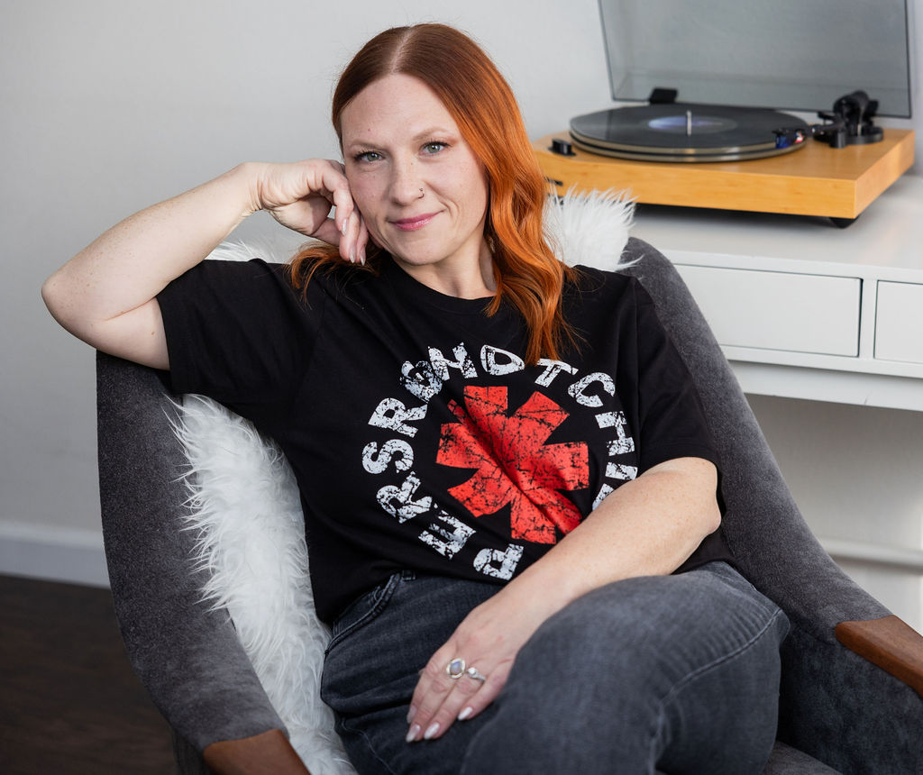 Small business bookkeeper sitting in a chair wearing a graphic band tee, with a record player in the background, representing a calm and modern approach to bookkeeping.